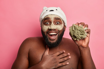 Headshot of Afro man has fun in bathroom, takes shower, holds sponge, applies clay mask on face for rejuvenation effect, smiles broadly, has white perfect teeth, stands topless against pink wall