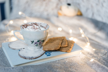 Hot chocolate and cookies laying on book in bed with lights, cozy winter concept