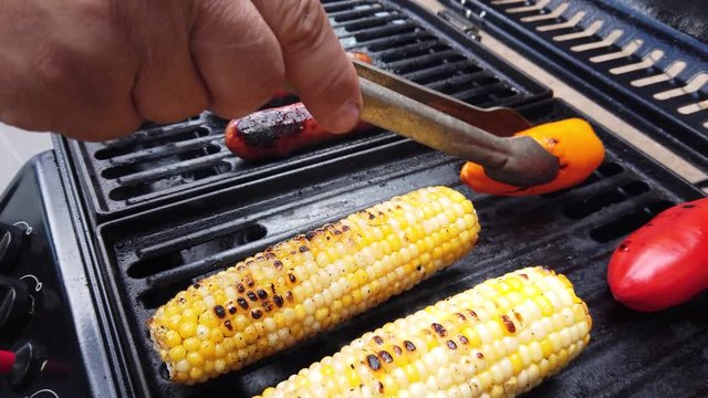 Man's hand turning bell peppers on a grill with corn and hot dogs