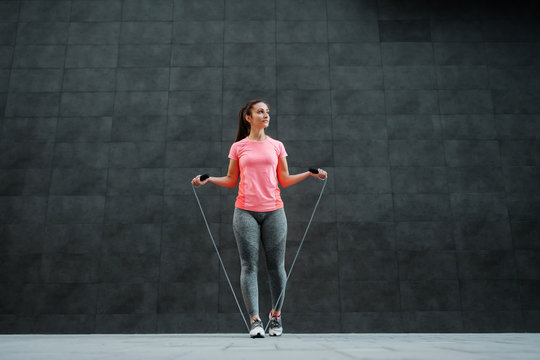 Full length of fit attractive caucasian sportswoman looking away and preparing to skip skipping rope. In background is dark wall.