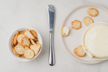 Cheese and cracker in a white bowl with a knife on a white background. Top view.