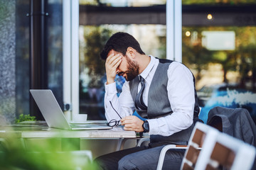 Nervous caucasian handsome bearded businessman in suit sitting in cafe and holding head. On table are laptop, coffee and eyeglasses. Trouble at work.