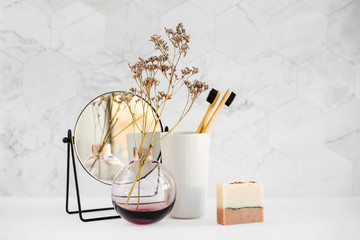 Branch in a glass vase, mirror, bamboo toothbrush and cosmetics products on white table. Decor for interior. Stylish decoration for home.
