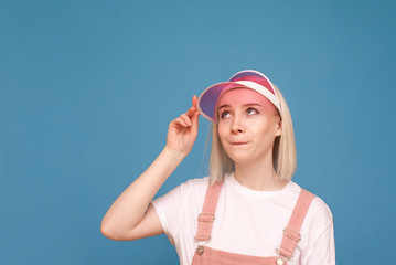 Fototapeta na wymiar Cheerful girl in cute clothes stands on a blue background, makes a funny face and looks away. Portrait of a funny girl with a pink cap on the background of the wall. Isolated.