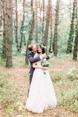 Happy and smiling wedding couple in the forest. Handsome groom embacing his pretty bride with floral wreath on the head and holding bouquet