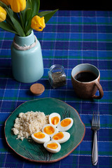 A healthy and nutritious breakfast: a cup of tea; a plate of hardboilde eggs with cuscus. A tulip bouquet in a vase, a checkered tablecloth on a shaded background.