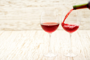 Red wine pouring into glasses from bottle, on a white wooden background