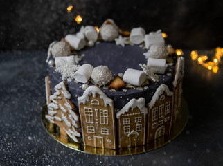 Obraz na płótnie Canvas Christmas gray cake decoraited of gingerbread cookies in shape of homes and snowy trees, marshmallows, snowflakes, sweets. Christmas, New Year greeting card. Gray background with garland. Copy space.