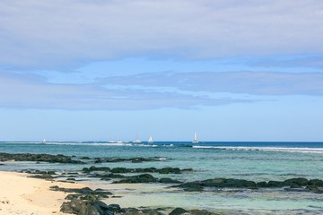 Mauritius coastline landscape.Typical scenic wild sand beach with black stones with coral reef on horizon and sailing boats and catamarans on background.Winter season on Mauritius.