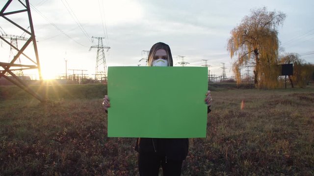 Young attractive girl in face mask holding empty blank green chromakey poster standing outside with sunset sky and moving cars on background. Air pollution. Global warming. Mockup. Free content.