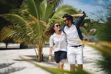 Beautiful young couple joyfully walks on the white sand on the seashore between green palm trees. Honeymoon Travel and Vacation on the Tropical Ocean