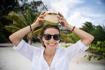 Young beautiful curly funny girl in a white shirt holds a coconut on her head, laughs, enjoys life, travel and vacation on the tropical ocean