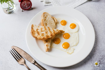 Breakfast time. Two fried eggs with toast for breakfast on a textured light gray background with a newspaper and an alarm clock