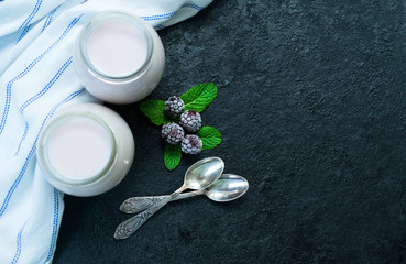 Blackberry yogurt in a jar and two small spoons on a black background. Copy space. Flat lay.