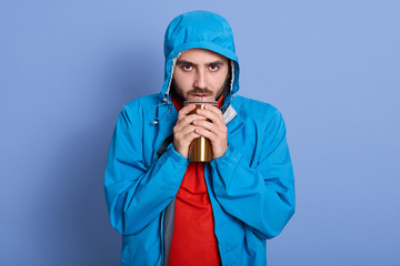 Studio shot of attractive young man with beard, having unhappy facial expression, holding thermo mug with coffee or tea, dresses blue jacket with hood and red shirt, being cold, basking hands.