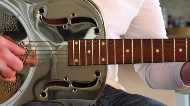 Man plays a resonator/national steel acoustic slide guitar in the kitchen's beautiful evening sunshine. Medium Close up with picking hand in shot.