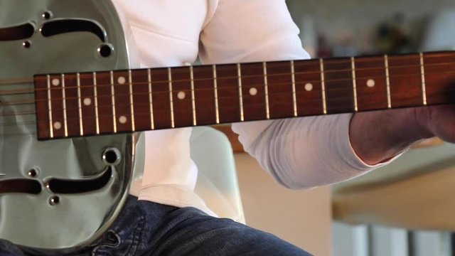 Man plays a resonator/national steel acoustic slide guitar in the kitchen's beautiful evening sunshine. Medium Close up.