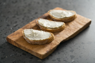 Three ciabatta slices with cream cheese on olive wood board on terrazzo surface