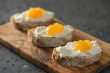 Three ciabatta slices with ricotta and orange marmalade on olive wood board on terrazzo surface