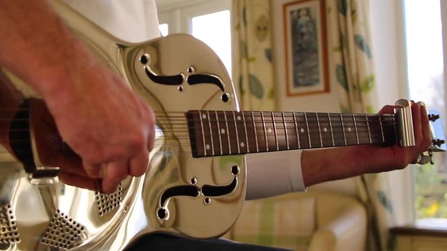 Man plays a resonator/national steel acoustic slide guitar in the kitchen's beautiful evening sunshine.