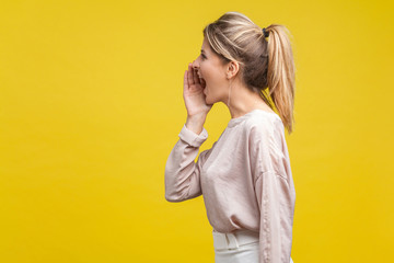 Profile side view of positive beautiful blonde woman with ponytale hairstyle and in casual beige blouse standing with hand over mouth and shouting. indoor studio shot isolated on yellow background