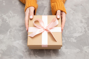 Beautiful female hands in warm yellow sweater holding gift box from kraft paper on gray concrete background, top view loft