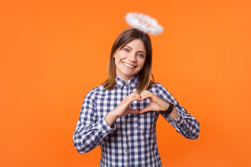 Portrait of angelic young woman with charming smile wearing checkered shirt standing with halo on head, showing heart sign with arms, charity and love. indoor studio shot isolated on orange background