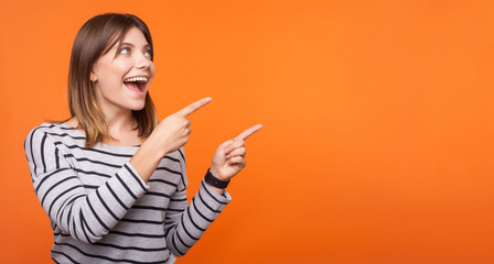 Portrait of surprised happy young woman with brown hair in striped shirt standing, pointing at empty space for announcement, unbelievable news. indoor studio shot isolated on orange background