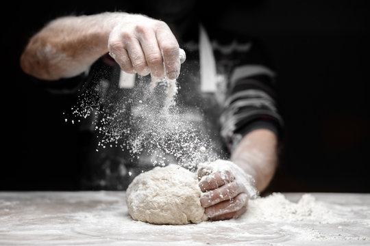 White flour flies in air on black background, pastry chef claps hands and prepares yeast dough for pizza pasta