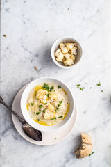 Roasted cauliflower cream soup on white kitchen table, served with fresh parsley, olive oil and bread. Overhead view.