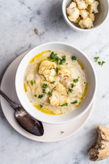 Roasted cauliflower cream soup on white kitchen table, served with fresh parsley, olive oil and bread. Overhead view.