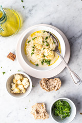 Roasted cauliflower cream soup on white kitchen table, served with fresh parsley, olive oil and bread. Overhead view.