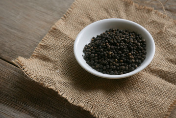 Black pepper in a white plate on rug sack on wooden background. 