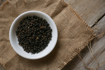 Black pepper in a white plate on rug sack on wooden background. 