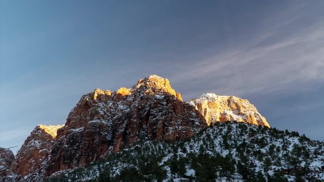 Timelapse of snow covered mountain top in Zion at sunset as thin clouds move through the sky and the shadow moves across the red rock cliffs glowing.