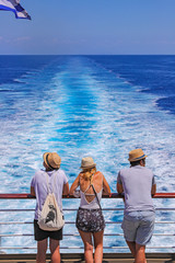 Tourists with a straw hat stand on the deck of a cruise ship and look out over the ocean  While the boat is sailing.