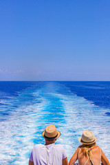 Tourists with a straw hat stand on the deck of a cruise ship and look out over the ocean  While the boat is sailing. Couple are looking at the sea as they stand at the edge of the ship's deck.