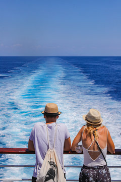 Tourists with a straw hat stand on the deck of a cruise ship and look out over the ocean  While the boat is sailing. Couple are looking at the sea as they stand at the edge of the ship's deck.