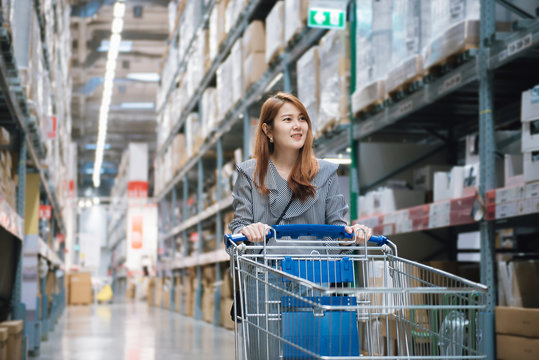 Young Asian Woman In Smiley With Cart Shopping In Furniture Warehouse Store.