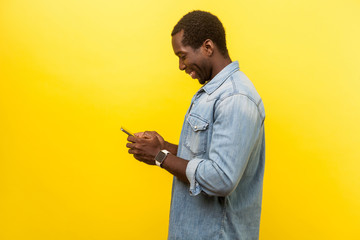 Profile of positive smiley man in denim casual shirt using cellphone, typing text message or...