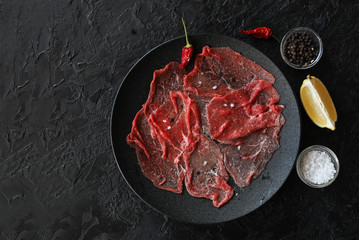 Marbled beef carpaccio with lemon, olive oil and Parmesan cheese on a black slate background. Top view, with copy space