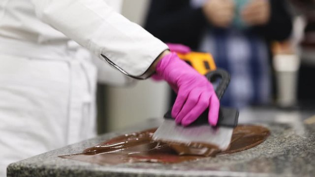 pastry chef in white uniform and pink gloves tempers chocolate on a marble plate using a scraper and pyrometer