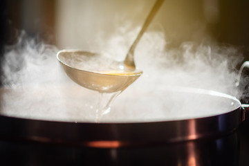 Cooking person in the restaurant is cooking while using the dipper in a large soup pot. And the water is boiling, there is smoke and hot spit.