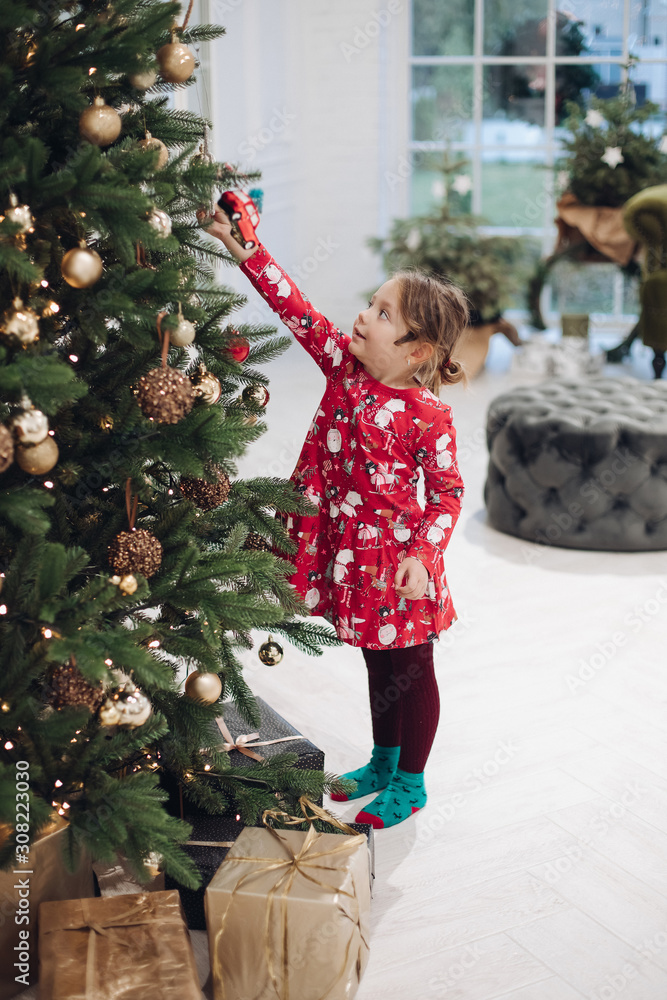 Wall mural Cute little girl in red dress touching toy ball on Christmas tree at luxury home interior long shot. Enthusiastic female baby enjoying New Year holiday surrounded by snowflakes having positive emotion