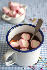Top view of cup of rustic chocolate with spoon and unfocused marshmallows in the background, on blue and white checkered tablecloth, in vertical