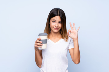 Fototapeta premium Young woman over isolated blue background holding coffee to take away while making OK sign