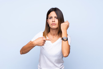 Young woman over isolated blue background showing the hand watch with serious expression serious because it is getting late