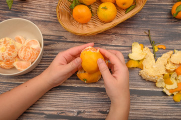 The girl's hands are cleaning tangerine, tangerines on a twig with green leaves, peeled tangerines in a ceramic dish, tangerine peel on a wooden background. Autumn harvest.