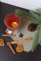 A glass of red drink stands on a piece of linen. A slice of dried orange and anise floats in it. Nearby are cinnamon sticks and dried oranges and a spruce branch.