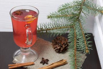 A glass of red drink stands on a piece of linen. A slice of dried orange and anise floats in it. Nearby are cinnamon sticks and dried oranges and a spruce branch.